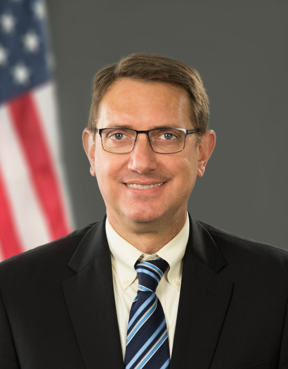 Professional headshot of a man with brown hair wearing glasses, a suit, and a blue-striped tie.